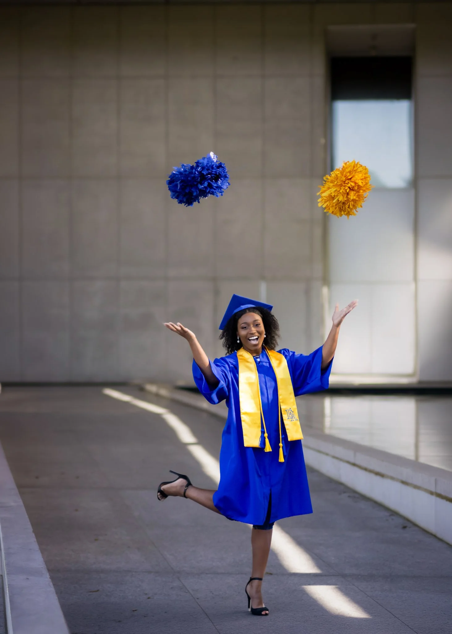 A joyful young woman in a blue graduation gown and cap, with a gold sash and stole, is celebrating outdoors. She is mid-jump with arms raised, smiling, and one leg bent at the knee. Pink and yellow pom-poms are flying in the air.