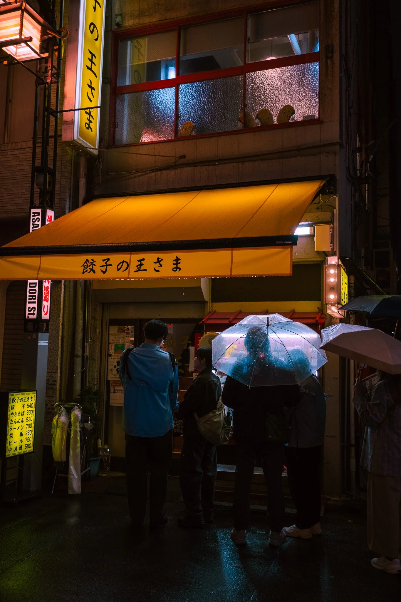 People with umbrellas outside a Tokyo ramen shop on a rainy evening.