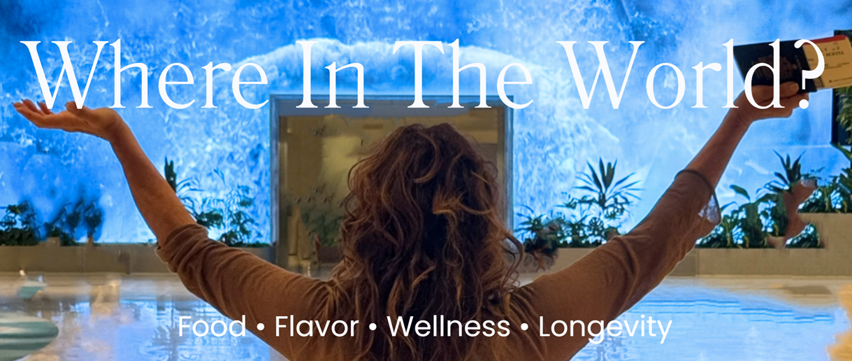 A woman with curly hair standing with her back to the camera, arms outstretched, in a spa-like setting with water and plants, looking at a large screen displaying ice and snow. Text reads "Where In The World?" and at the bottom, "Food, Flavor, Wellness, Longevity."