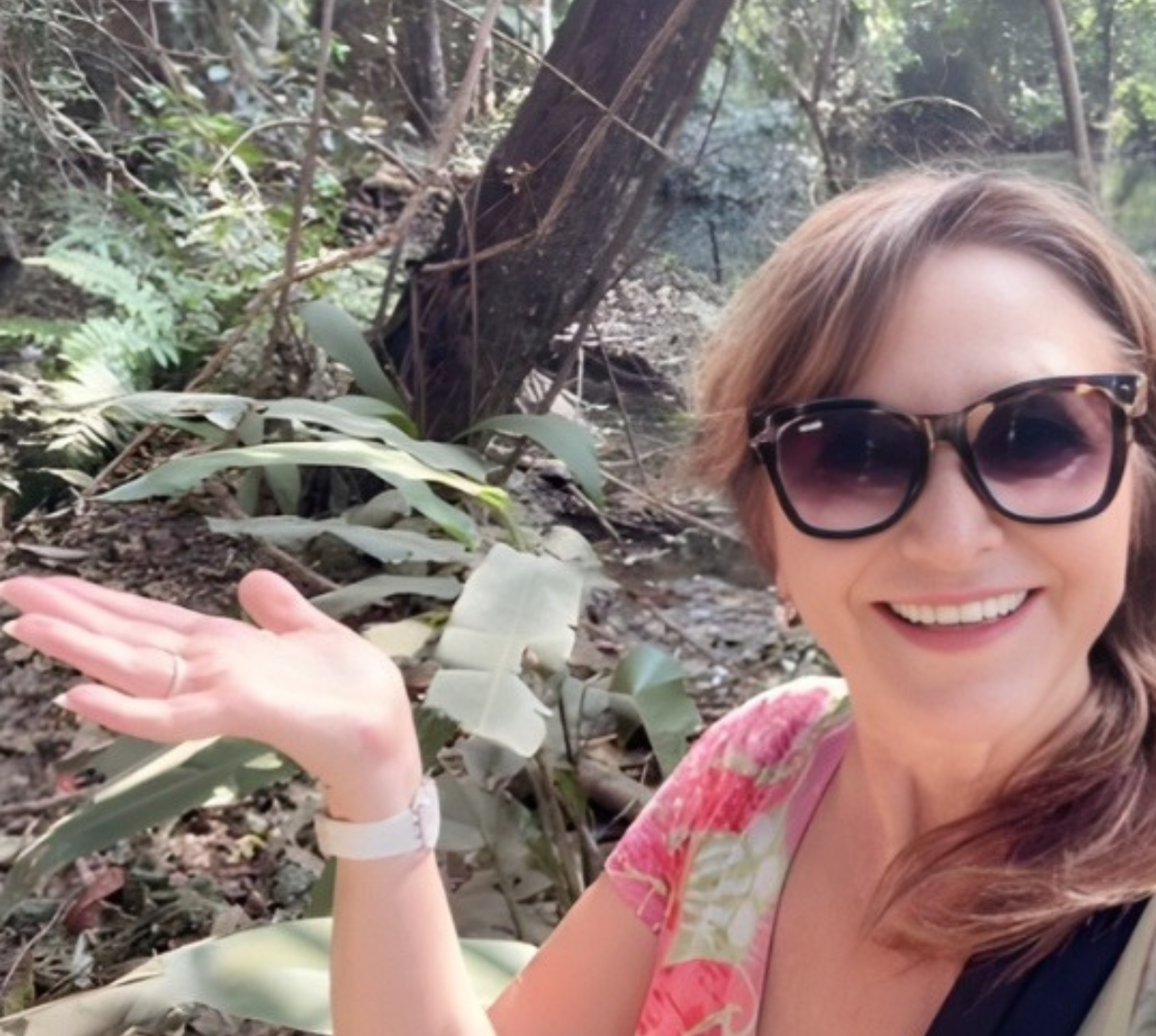 A woman with sunglasses, smiling, wearing a pink floral shirt, standing in a forest near a small stream with green plants and trees in the background.