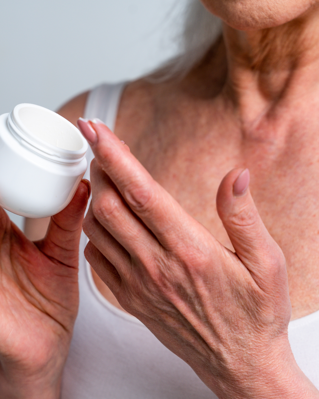 Close-up of an elderly woman applying lotion or cream from a white jar to her chest.