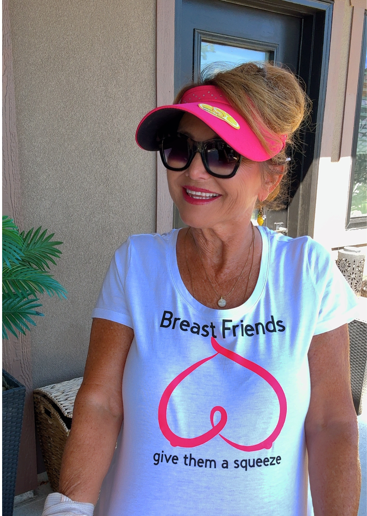 A smiling woman wearing a pink visor, dark sunglasses, and a white T-shirt that reads "Breast Friends" with a pink bra illustration and the phrase "give them a squeeze." She has earrings, a necklace, and appears to be sitting outdoors near a door and some plants.