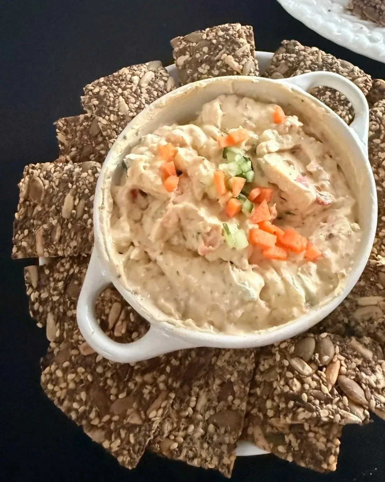 A white bowl of chicken and vegetable dip garnished with chopped carrots and celery, surrounded by sesame seed crackers on a black surface. Fiber Rich Seed crackers with a Oh So Bacon Dip
