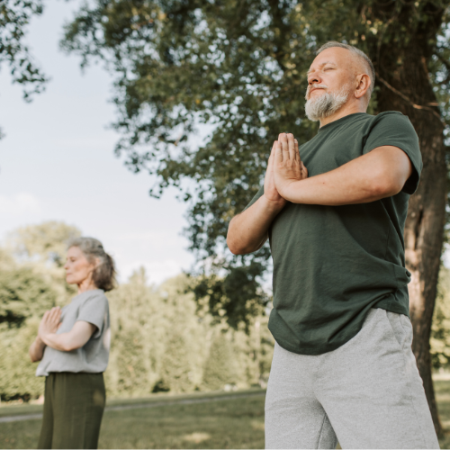 An older man and woman practicing yoga outdoors in a park, standing with their eyes closed and hands in a prayer position.