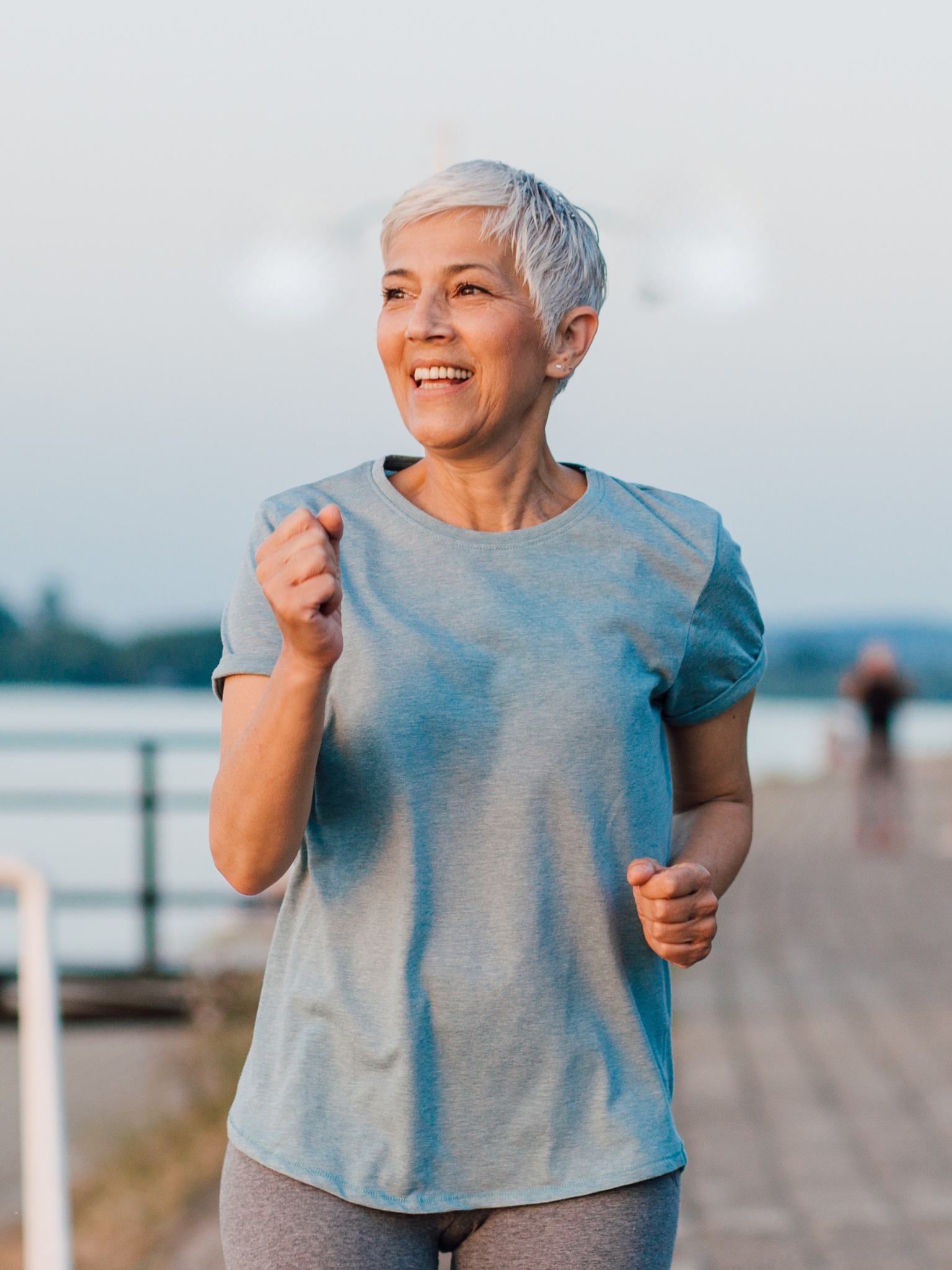 A woman jogging outdoors, smiling, wearing a light blue t-shirt and gray leggings, with a blurred background of water and boats.
