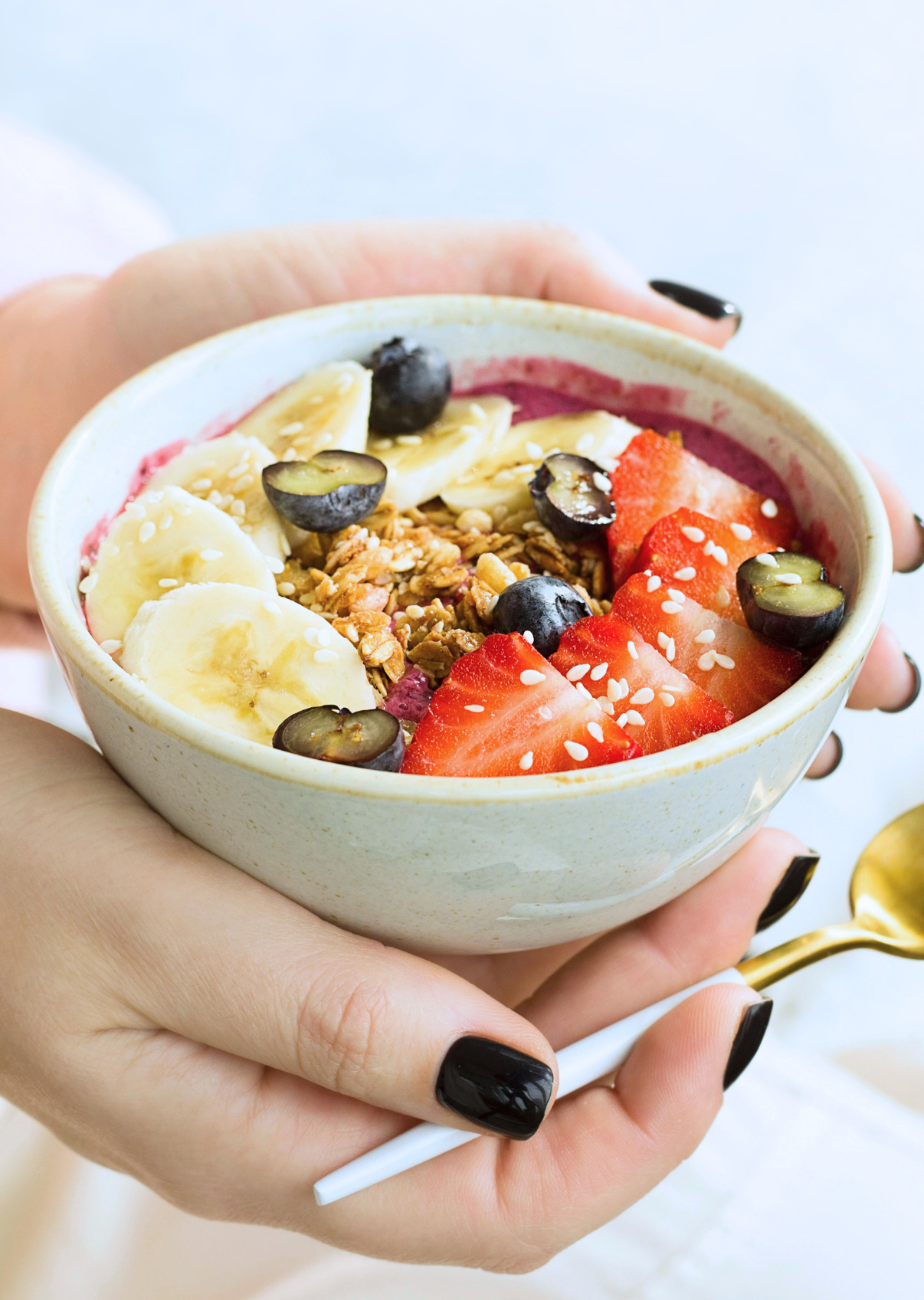 A hand holding a bowl of yogurt topped with sliced bananas, strawberries, blueberries, granola, and sesame seeds.