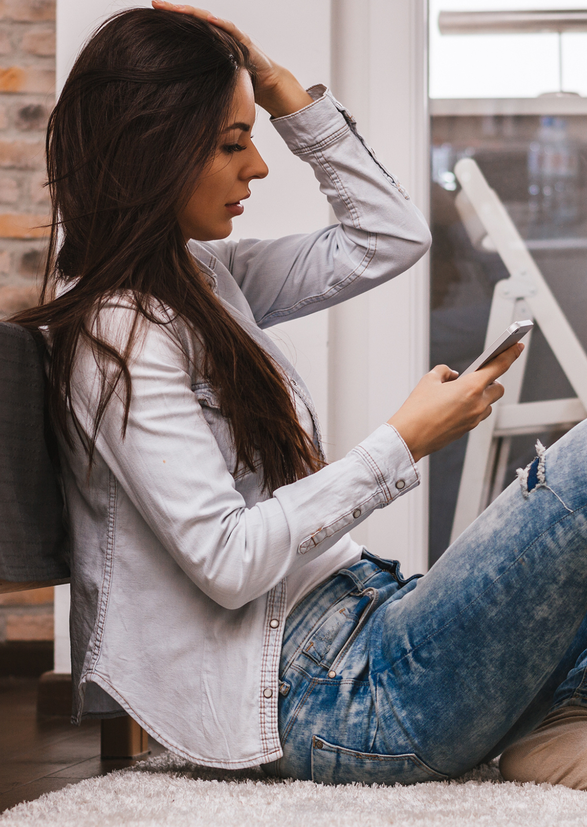 A woman sitting on the floor, leaning against a chair, looking at her phone with one hand on her head, in a room with a brick wall and window.