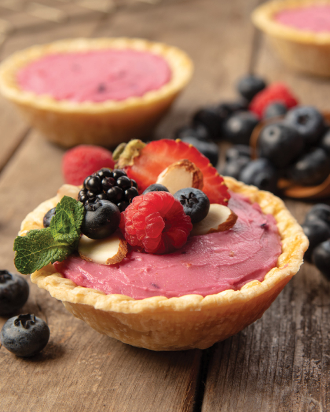 A tart filled with pink berry-flavored cream topped with fresh mixed berries and mint leaves, with additional berries scattered around on a wooden surface.