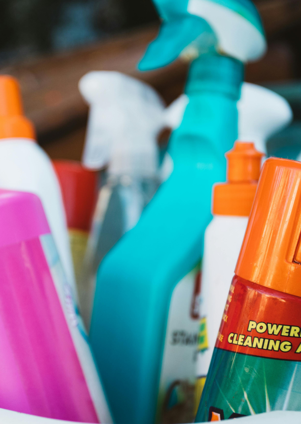 Close-up of various cleaning spray bottles with colorful caps and labels, including blue, orange, pink, and white bottles, some labeled with cleaning product descriptions.