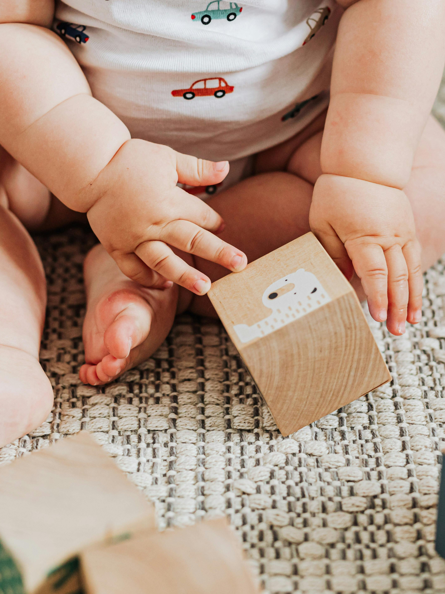 Close-up of a baby's hands and legs, wearing a white shirt with small colorful cars, playing with wooden blocks on a woven, textured surface.