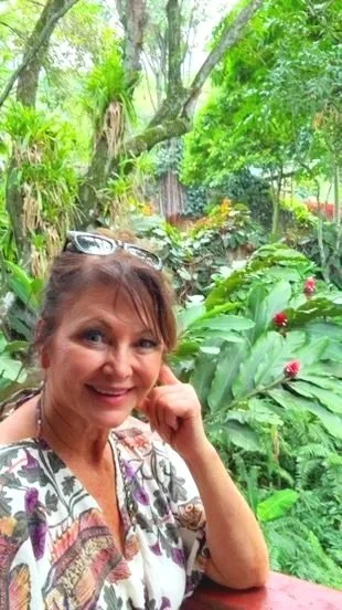 A woman smiling and resting her chin on her hand in a lush, green garden with various plants and red flowers.