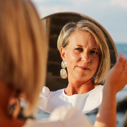 A woman looking at herself in a mirror, outdoors, with the ocean in the background.