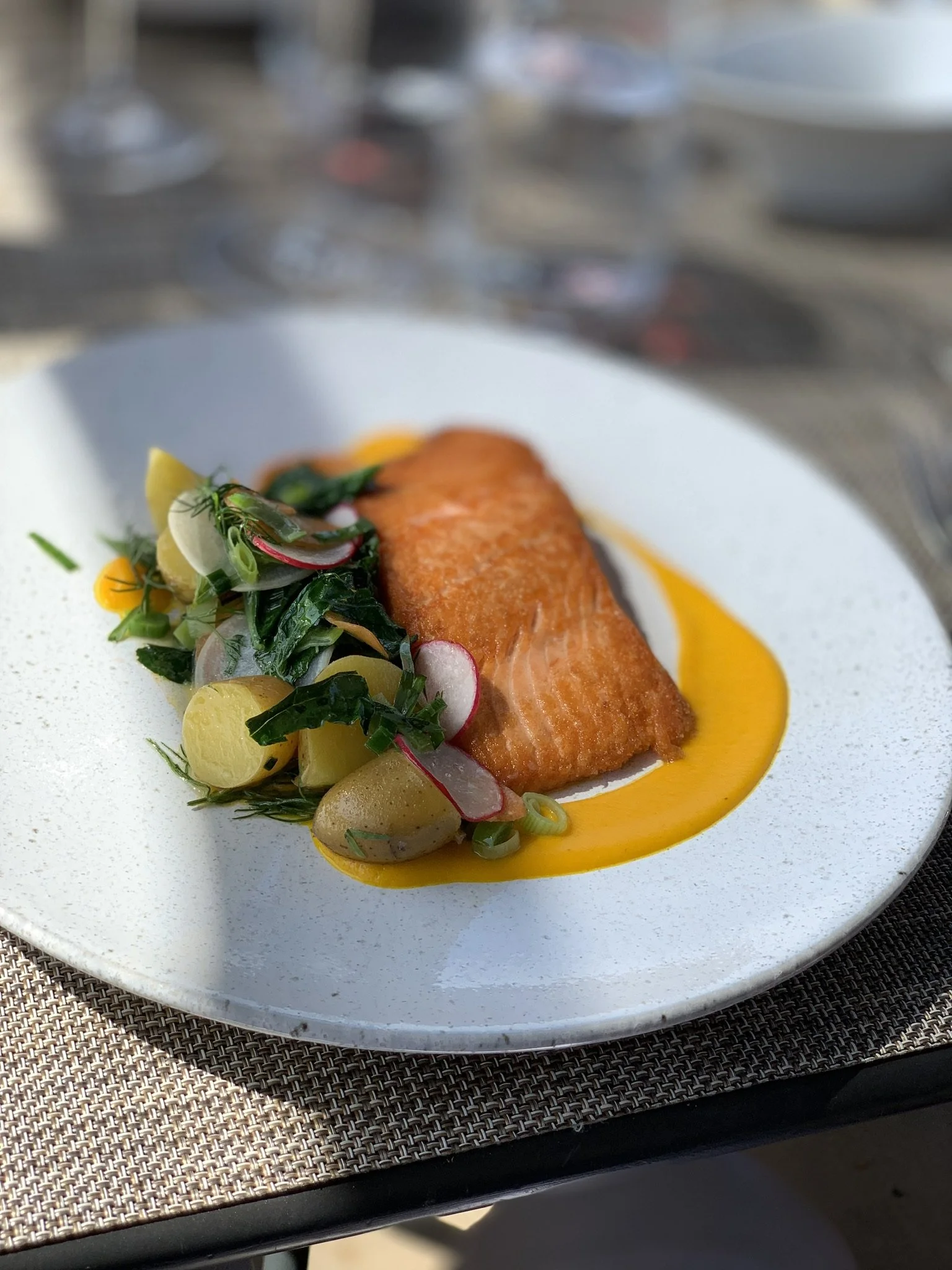 A plated dish featuring a piece of cooked fish fillet garnished with fresh herbs and radish, served with a vibrant yellow sauce on a white plate placed on a textured placemat.