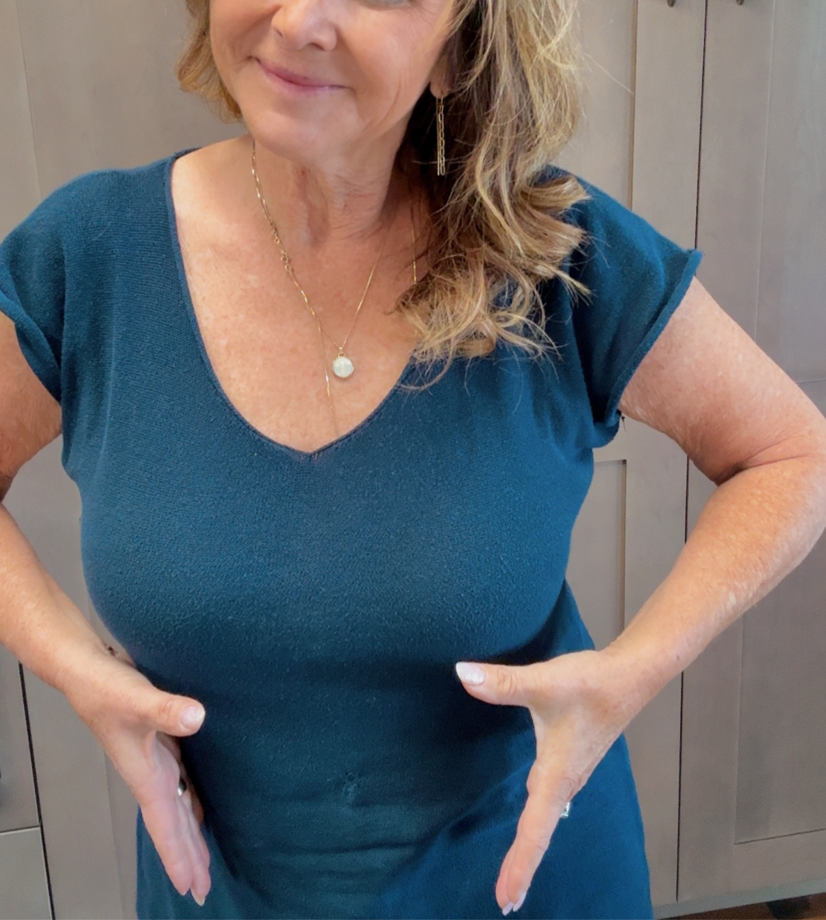 A woman with wavy blonde hair is wearing a blue V-neck T-shirt, a gold necklace with a white pendant, and matching gold earrings. She is smiling slightly and has her hands on her hips. The background features wooden panels.