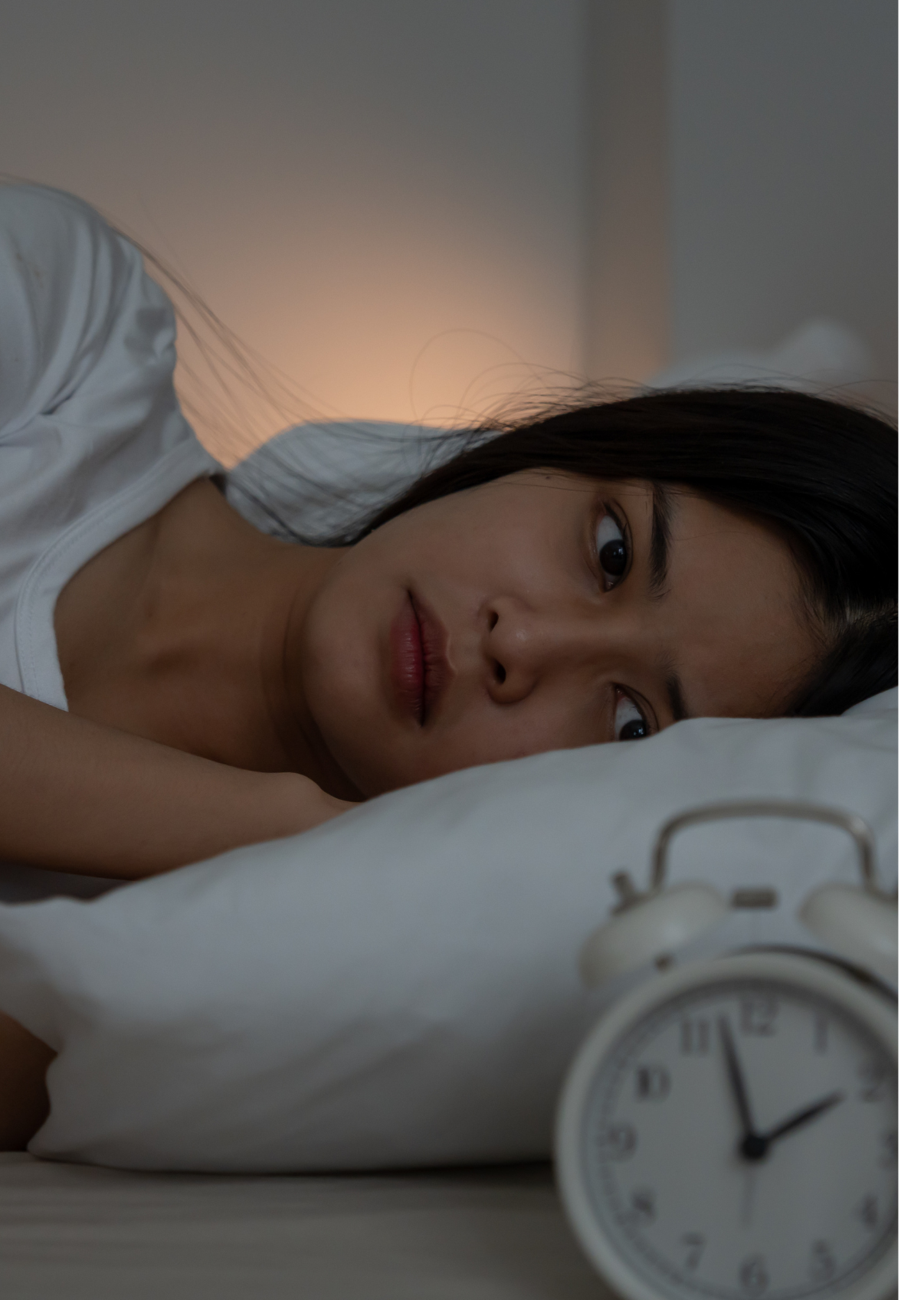 A young woman lying in bed, looking distressed, with an alarm clock showing 3:55 am on the bedside table.