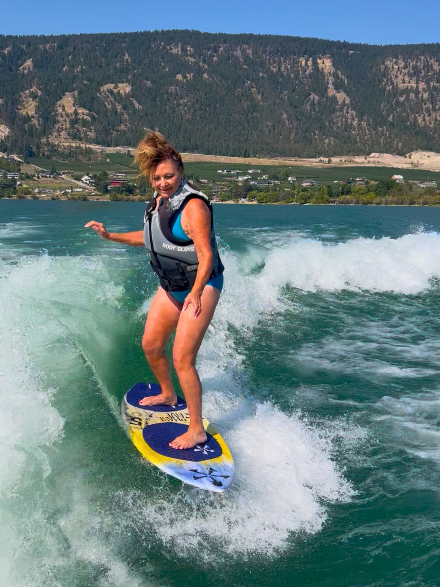 A woman surfing on a lake, wearing a life jacket, with mountains and a small town in the background.