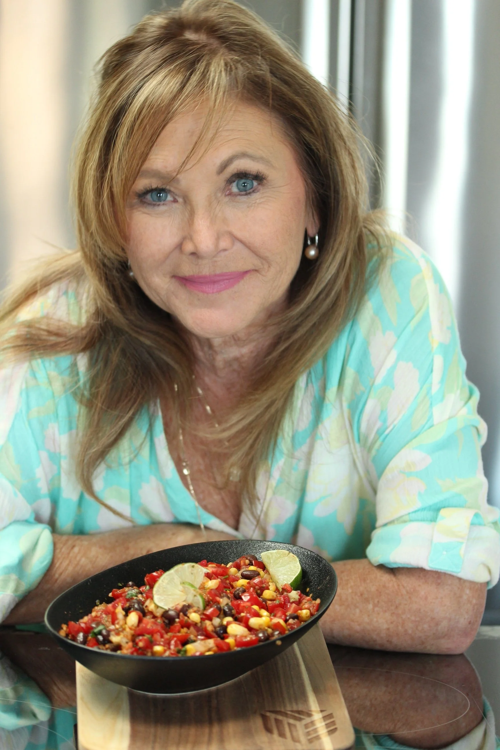 A woman with light brown hair, blue eyes, and pearl earrings is smiling at the camera. She is wearing a light blue and yellow patterned top. In front of her, there is a black bowl of colorful food, possibly salad or ceviche, garnished with lime wedges. The background shows a metallic surface, possibly a kitchen or restaurant setting.