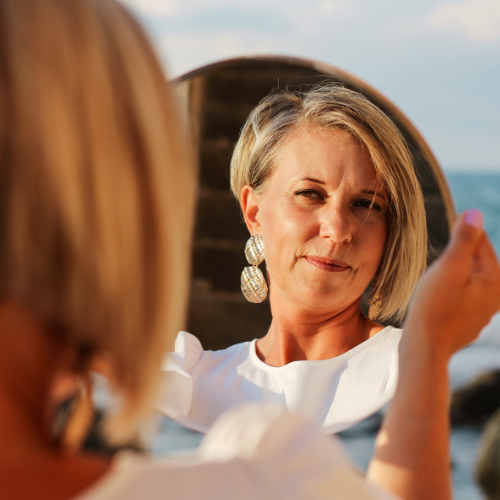 Woman with short blonde hair and earrings smiling at herself in a round mirror outdoors near the water.