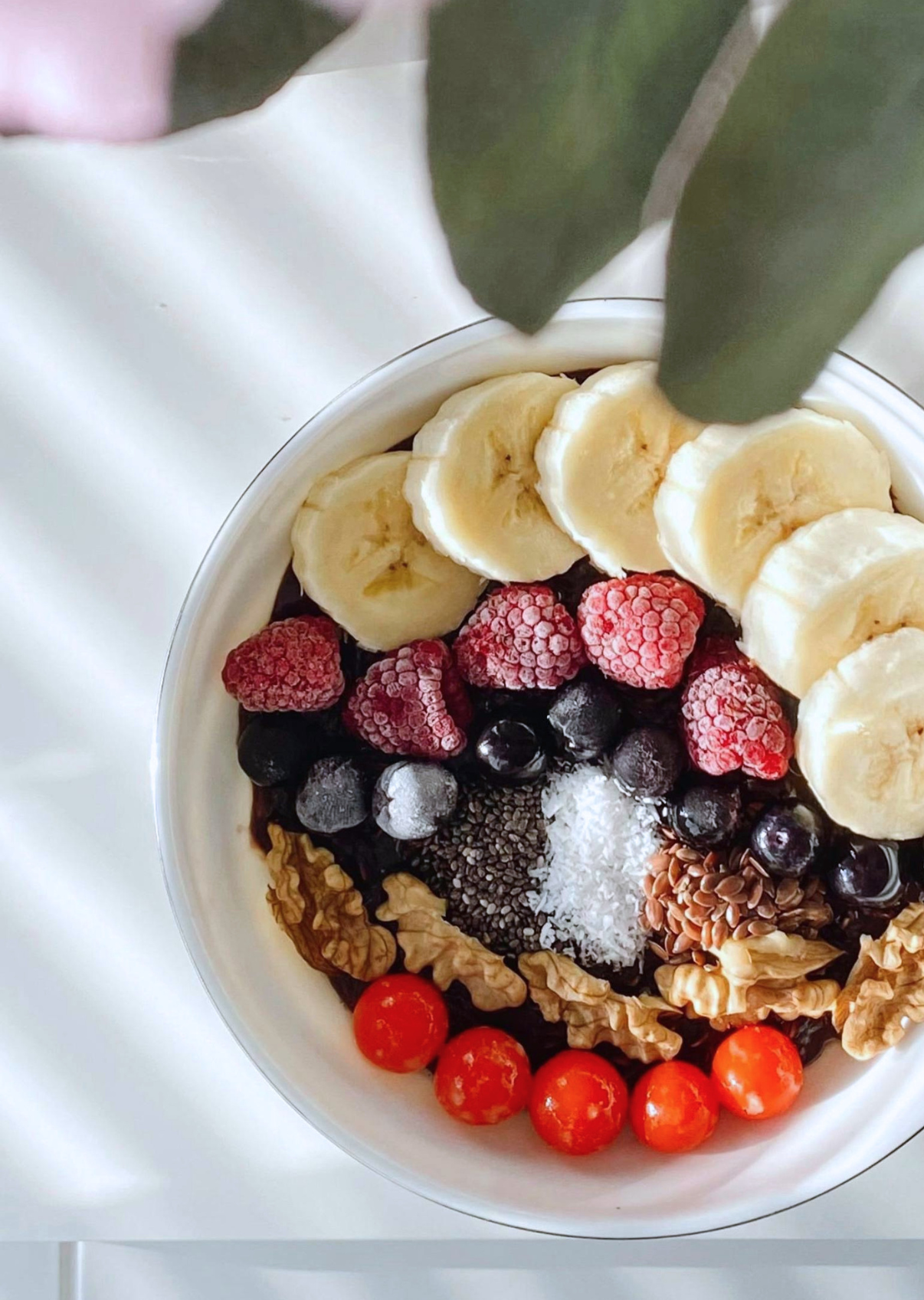 A bowl of fruit and toppings, including sliced bananas, red and black berries, cherry tomatoes, shredded coconut, chia seeds, flax seeds, and walnuts, on a white surface with a white cloth in the background.