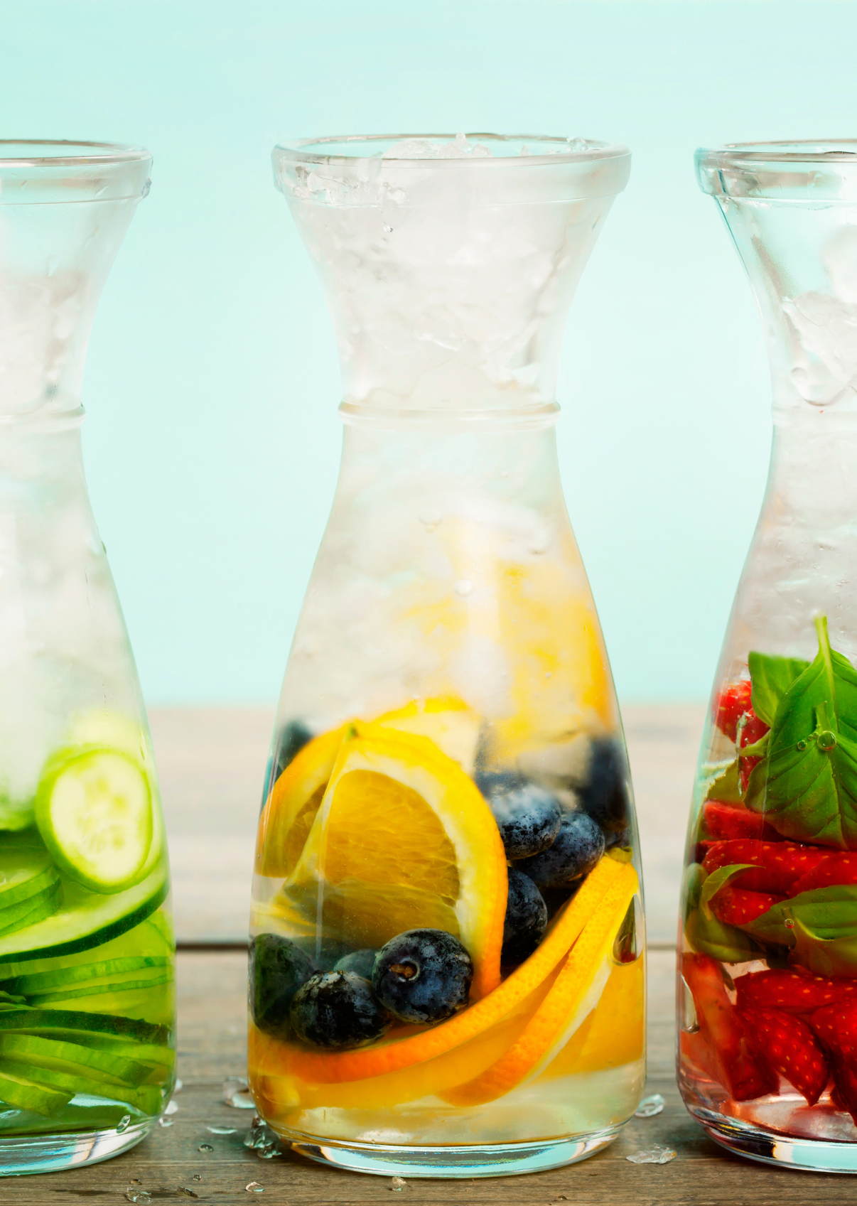 Three glass bottles containing infused water with slices of cucumber, orange, blueberry, strawberry, and fresh herbs on a wooden surface with a pastel blue background.