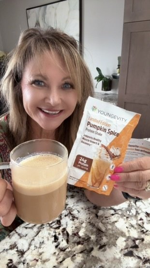 Woman smiling at the camera holding a glass of pumpkin spice protein shake and a box of Youngeity Pumpkin Spice Protein Shake powder in a kitchen.