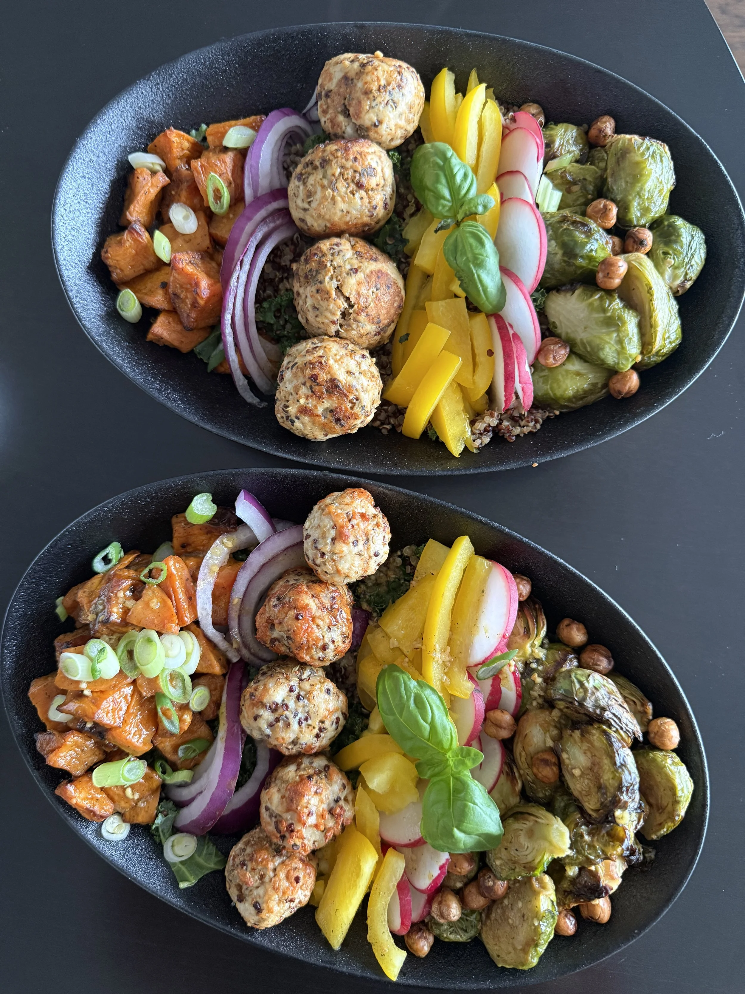 A black oval plate filled with colorful roasted vegetables, including sweet potatoes, Brussels sprouts, radish slices, yellow bell peppers, and cherry tomatoes, garnished with fresh basil leaves and chopped green onions.