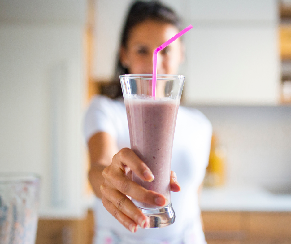 A woman holding a glass of pink smoothie with a pink straw, blurred background of a kitchen.
