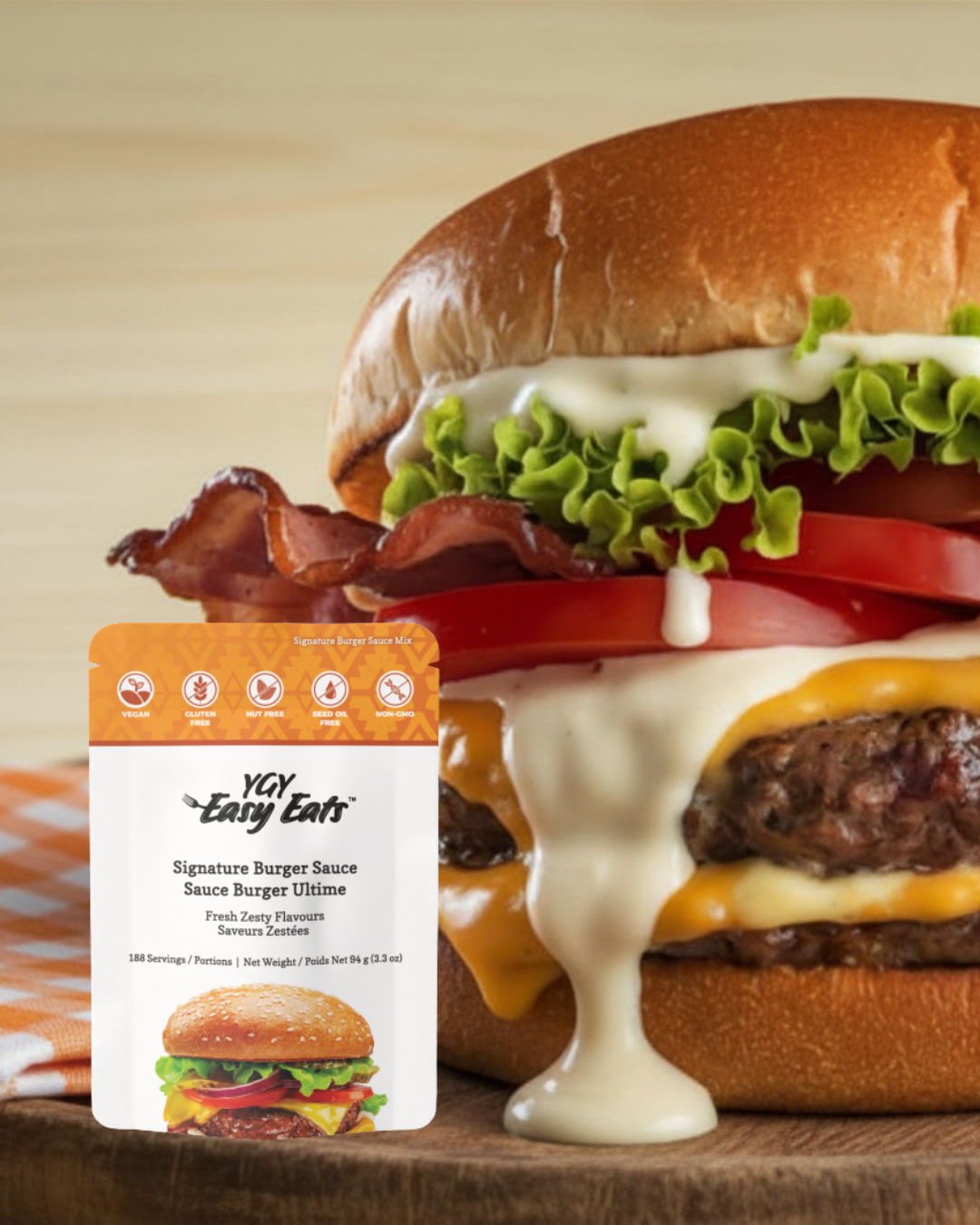 A close-up of a cheeseburger with lettuce, tomato, bacon, cheese, and a beef patty in a bun, with a packet of YAY Easy Eats Signature Burger Sauce in front.