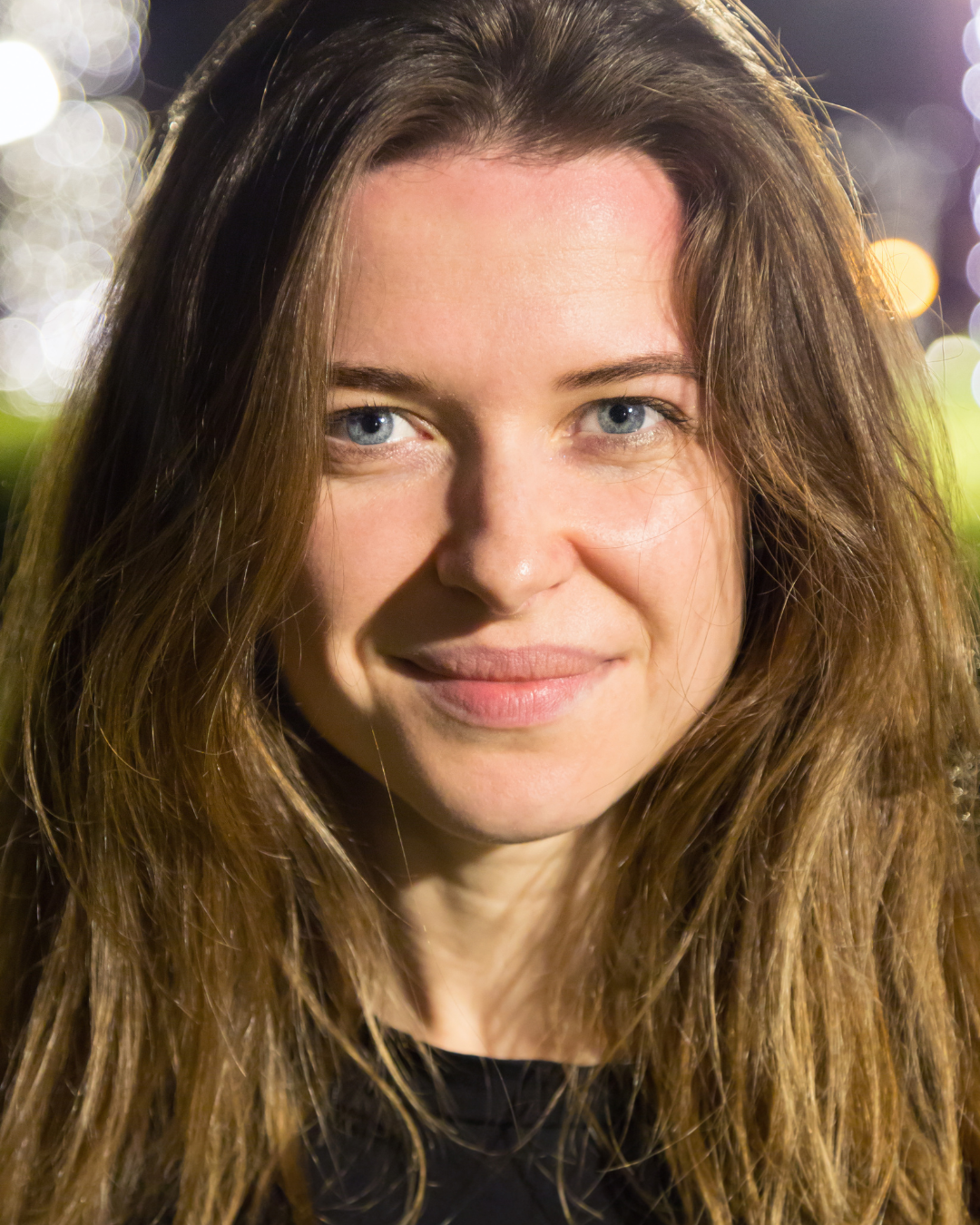 Close-up portrait of a woman with long, wavy brown hair and blue eyes, smiling at night with blurred lights in the background.