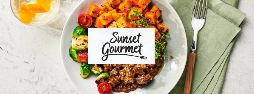 A white plate with roasted sweet potatoes, cherry tomatoes, and a vegetable salad, with a label reading "Sunset Gourmet" on top. A fork is on a green napkin, and a glass of water with lemon is in the background.