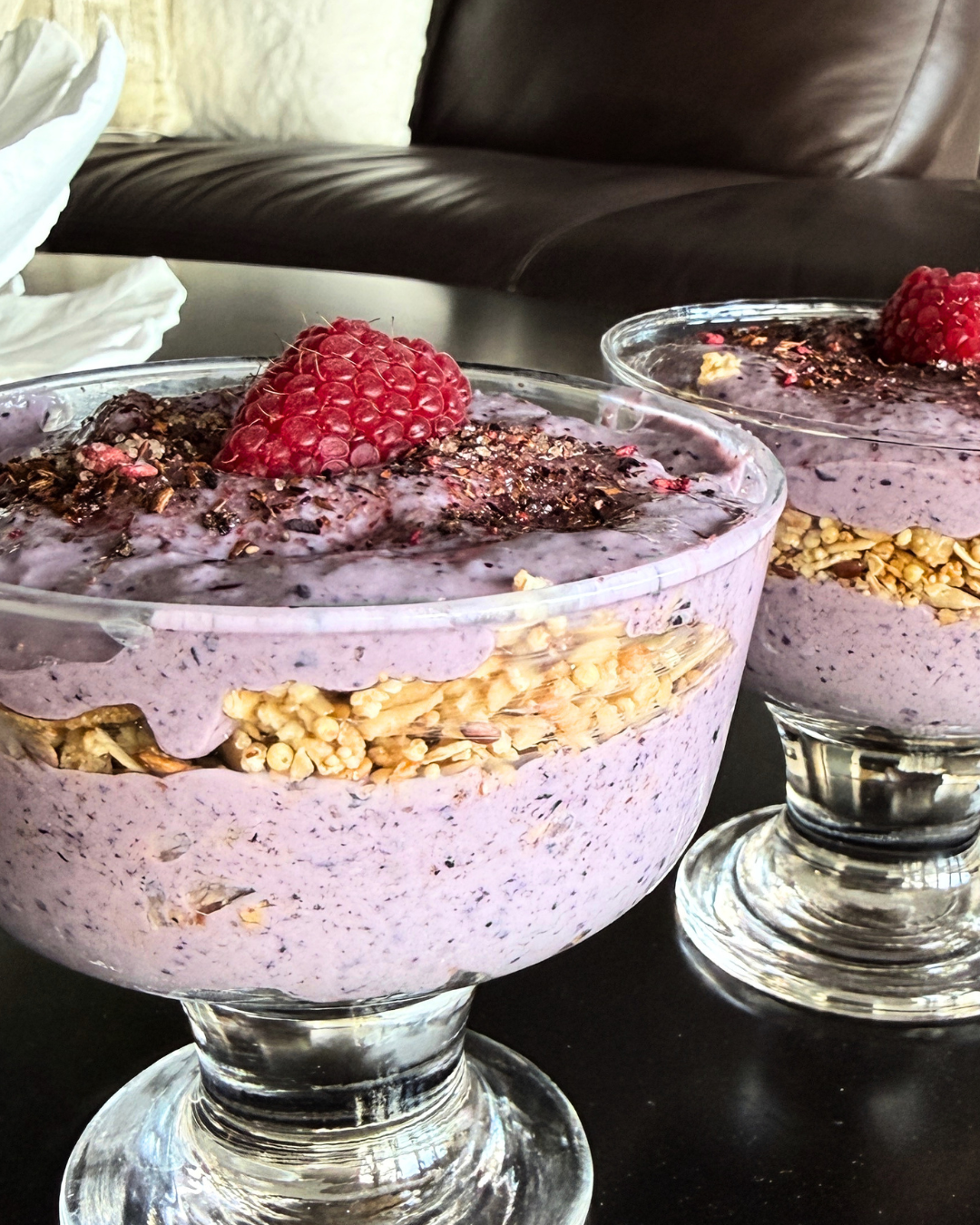 Two glass dessert cups filled with a layered berry yogurt parfait topped with a raspberry, served on a black table in front of a leather couch.