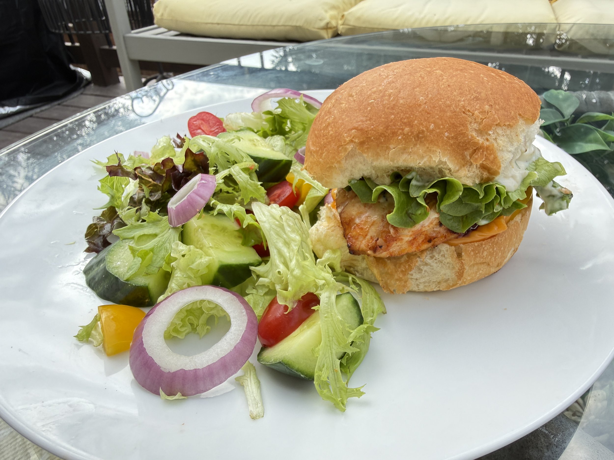 A close-up of a sandwich on a bun with lettuce, tomato, and possibly chicken, served alongside a salad with cucumbers, radishes, lettuce, and bell peppers on a white plate.