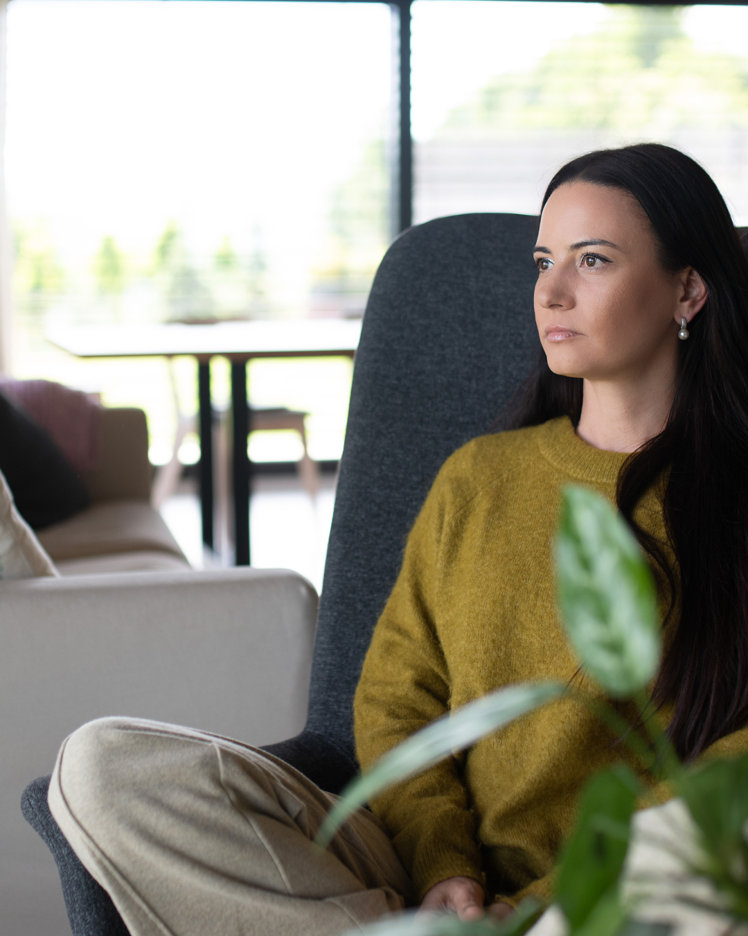 A woman with long dark hair and pearl earrings sitting calmly in a modern room, wearing a mustard sweater, with a large window and greenery outside.