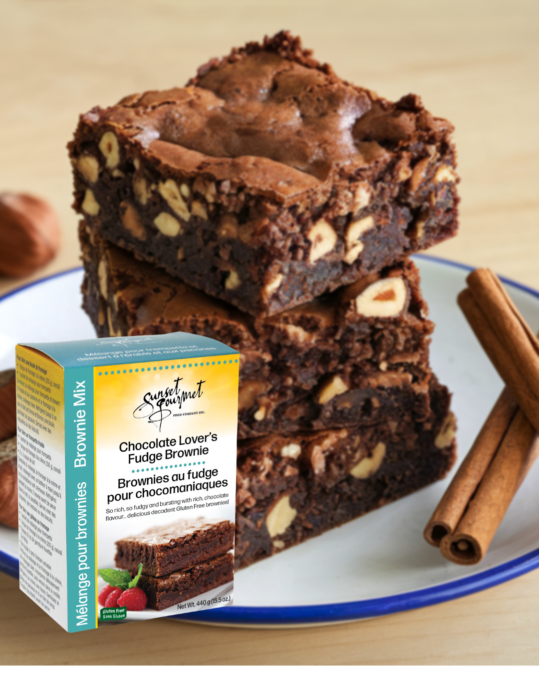 Stacked chocolate fudge brownies with chopped nuts on a white plate, cinnamon sticks nearby, and a box of Sunset Gourmet Brownie Mix in front.