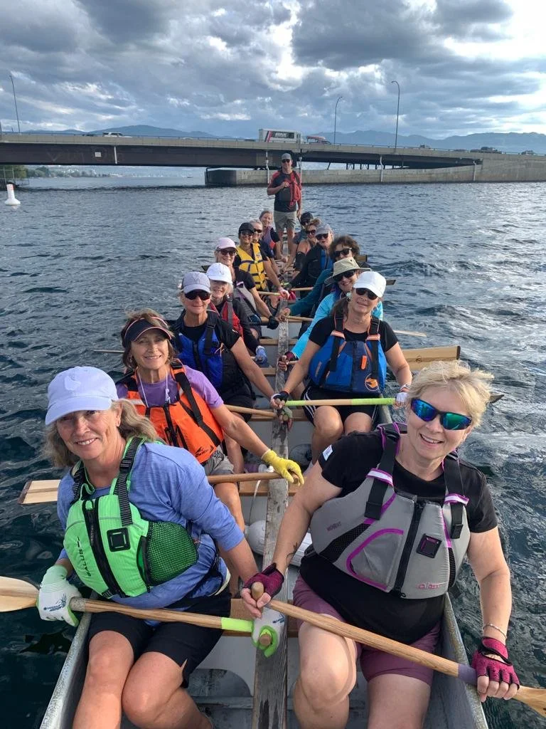 Group of women and one man paddling a dragon boat on a river on a cloudy day, wearing life jackets and sunglasses.