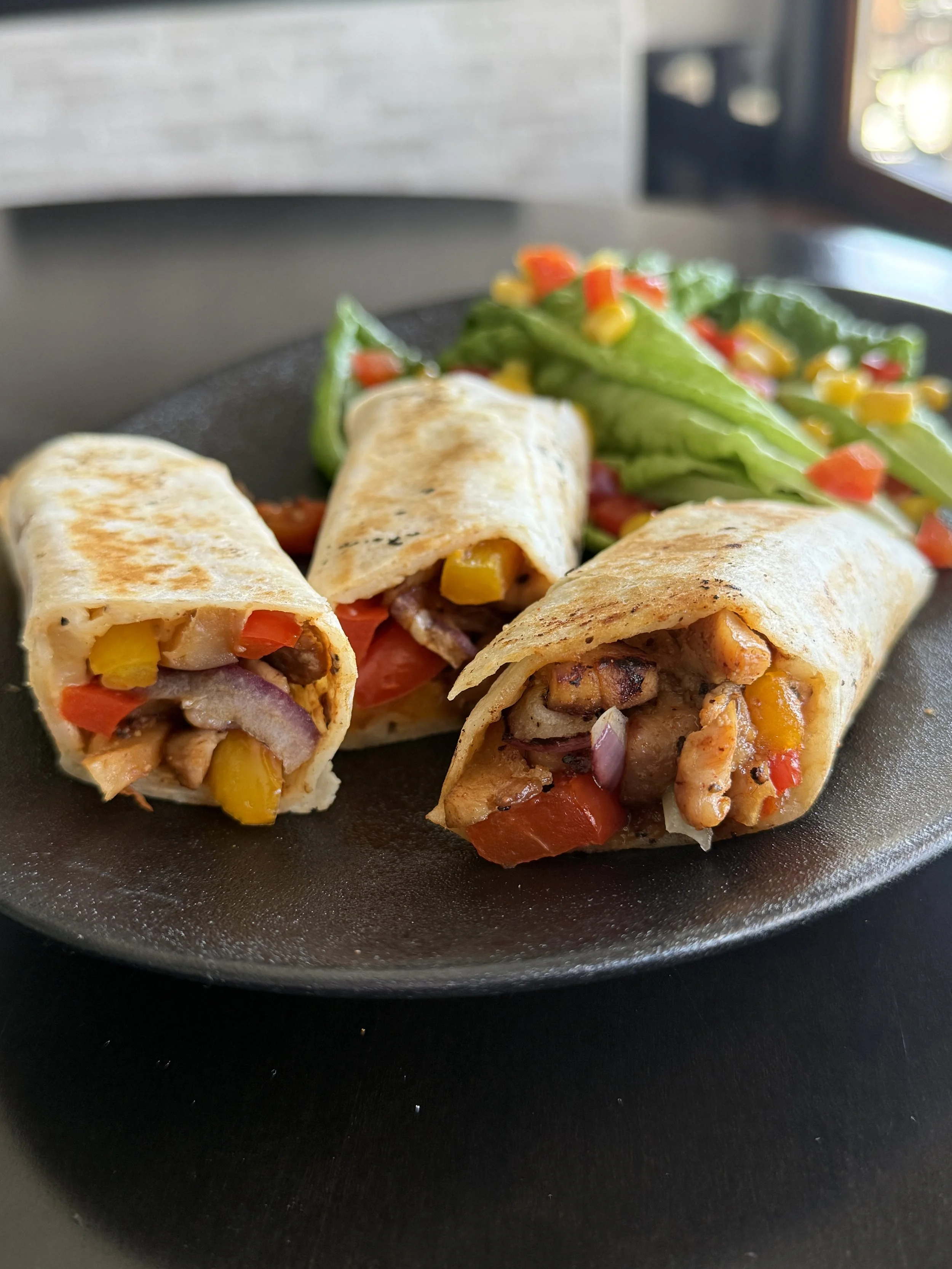 Close-up of three grilled  Sweet Maple Chicken Wraps with a side of mixed vegetables on a black plate.