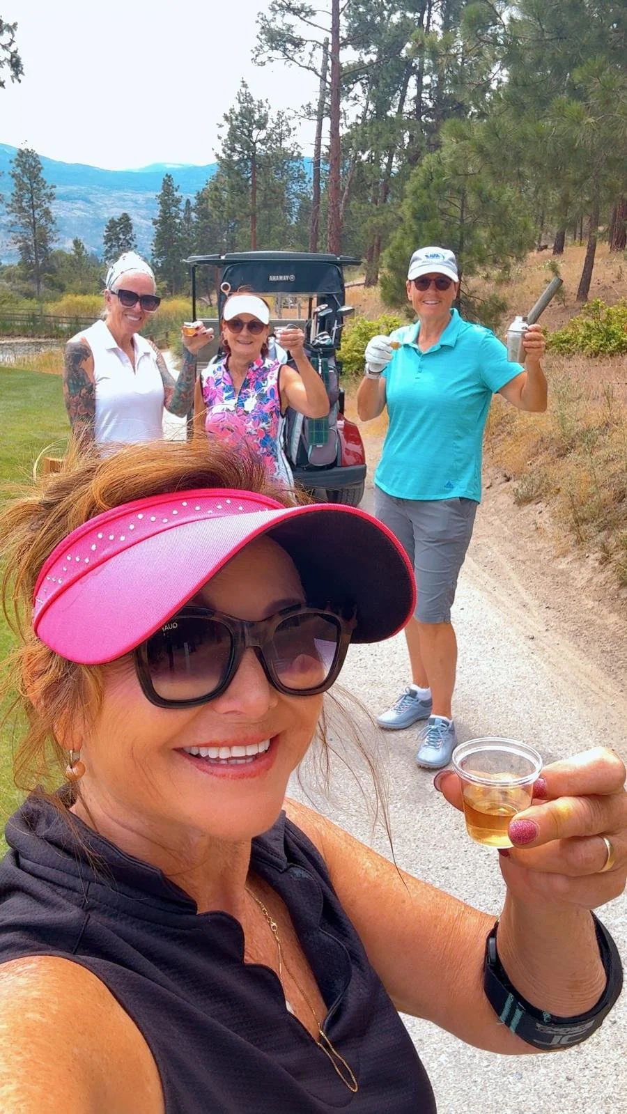 A group of five women on a golf course, smiling and enjoying their time. The woman in the foreground is taking a selfie, holding a small glass of what appears to be whiskey or another beverage, and is wearing a pink visor and sunglasses. Three women behind her are standing next to a golf cart, with one woman wearing a turquoise shirt and holding a pipe, smiling. The background features trees, mountains, and a partly cloudy sky.