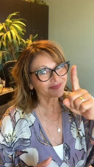 A woman with glasses and shoulder-length hair pointing her index finger upward. She is indoors with a potted plant and dark furniture in the background.