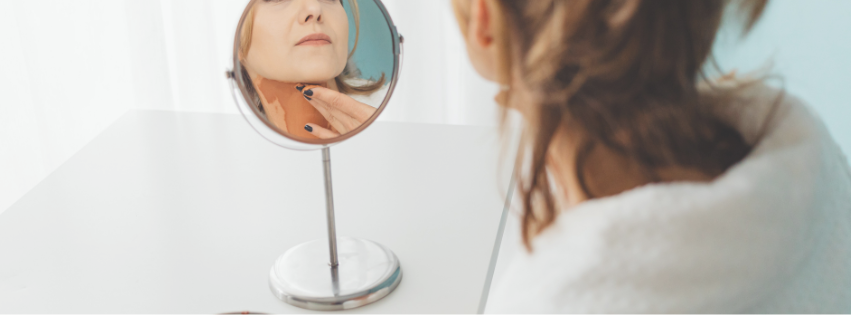 Woman looking at her reflection in a mirror from behind, with her face and makeup visible in the mirror.