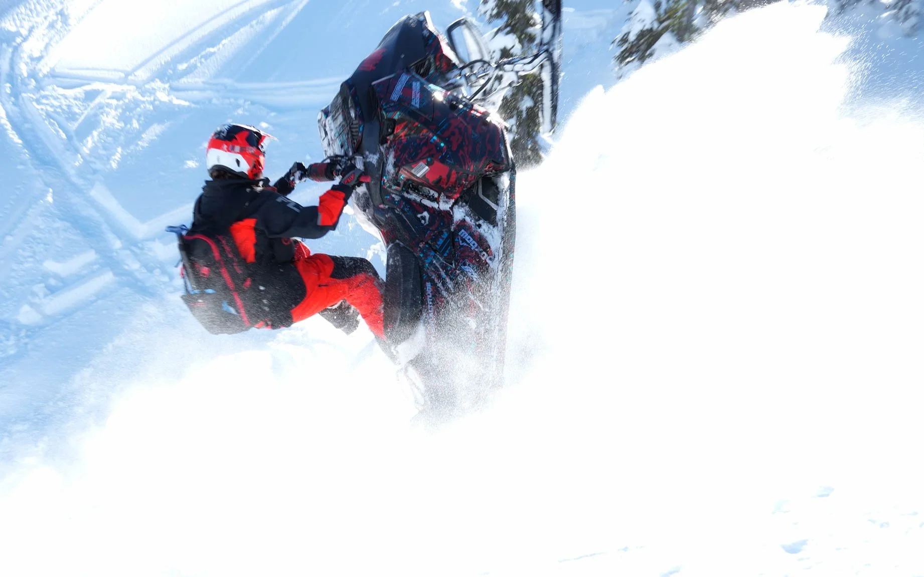 Person in black and red winter gear riding a snowmobile through snowy terrain with trees in the background.