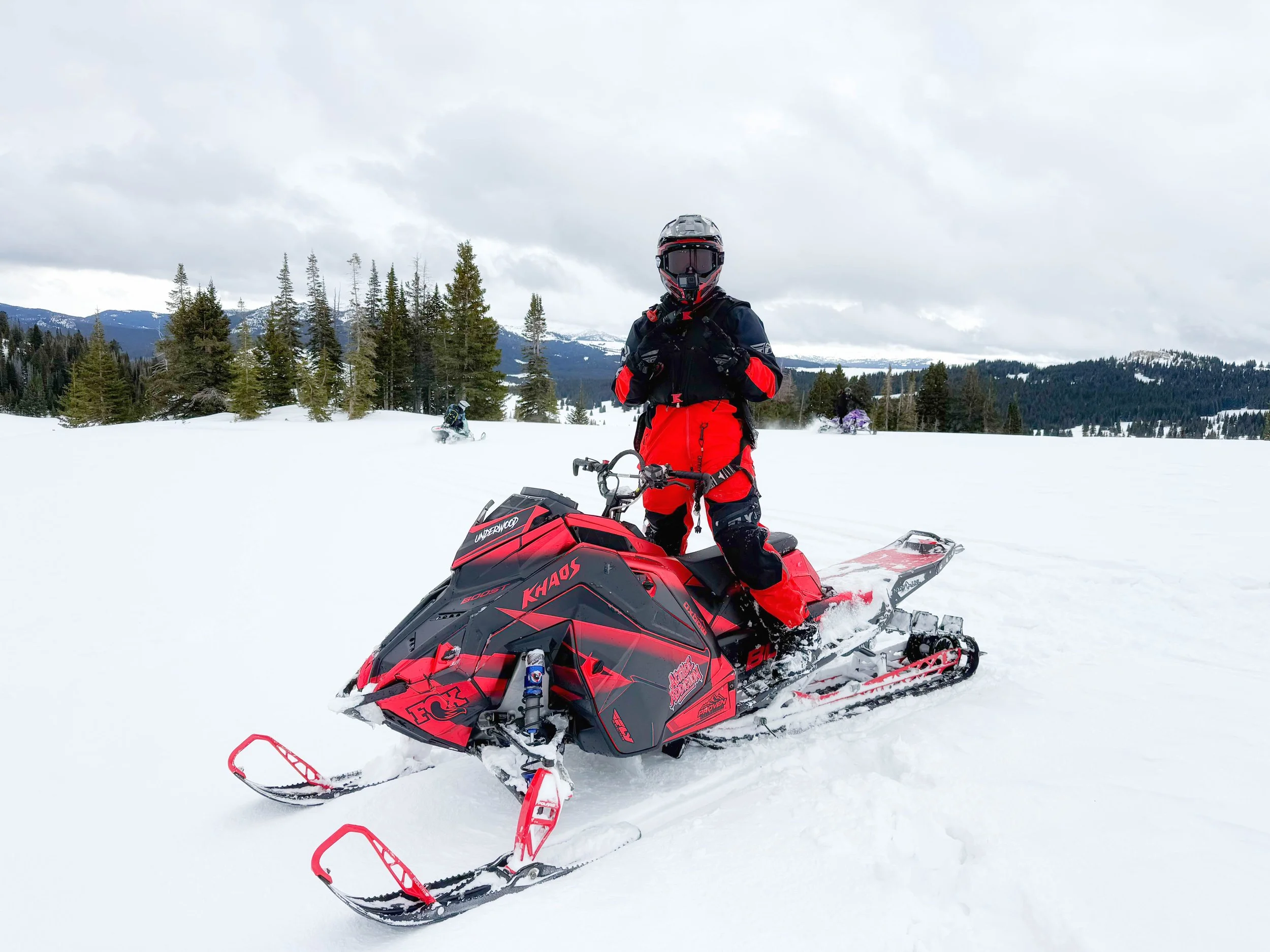 A person dressed in black and red winter gear standing next to a red snowmobile on a snowy landscape with pine trees and mountains in the background.