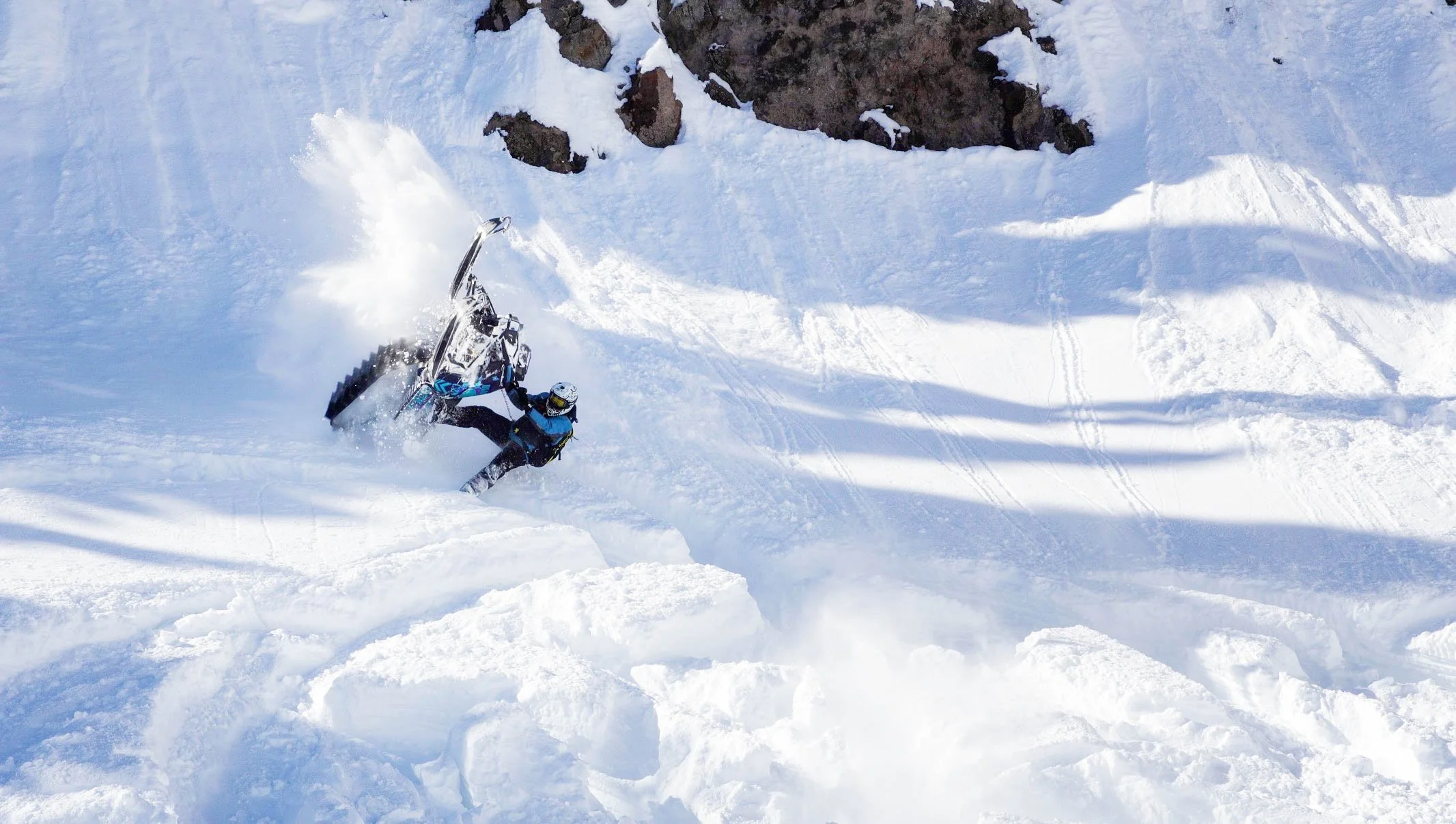 An individual falling while snowboarding on a snowy slope with rocks in the background.