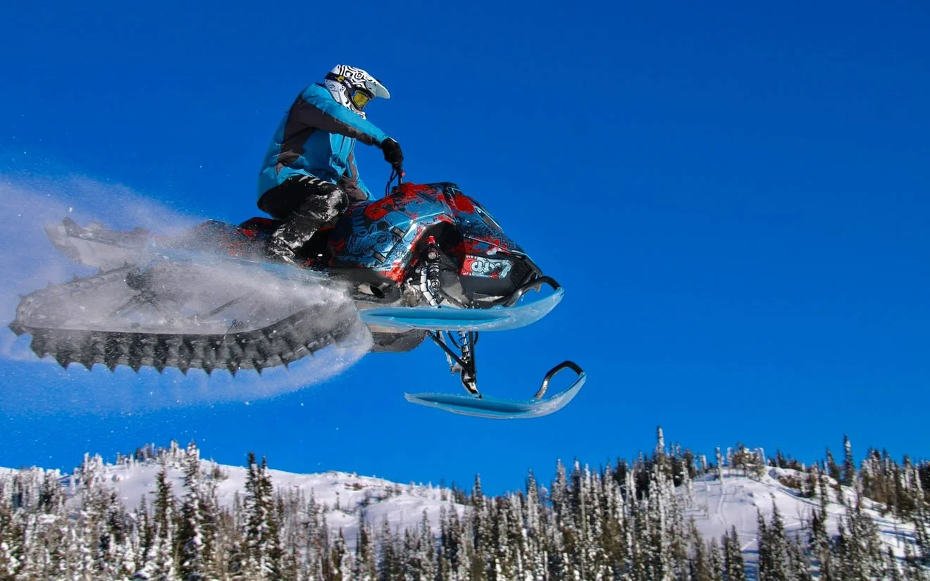 A person riding a snowmobile mid-air over a snowy landscape with snow-covered trees and a clear blue sky.