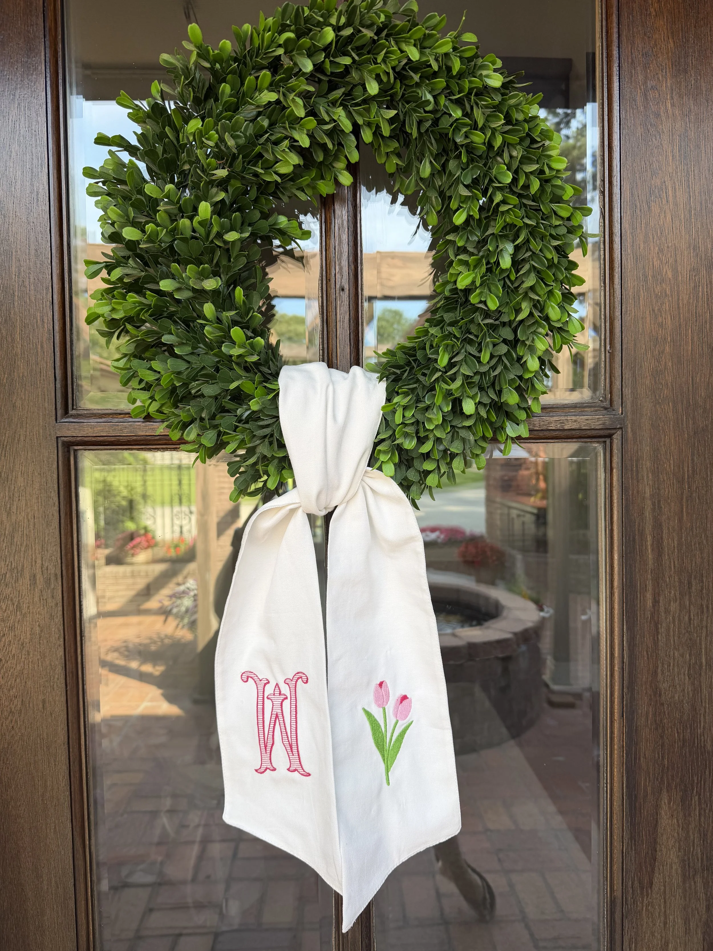 Decorative greenery wreath with a white cloth tied in a bow at the bottom, hanging on a wooden door with glass panels.