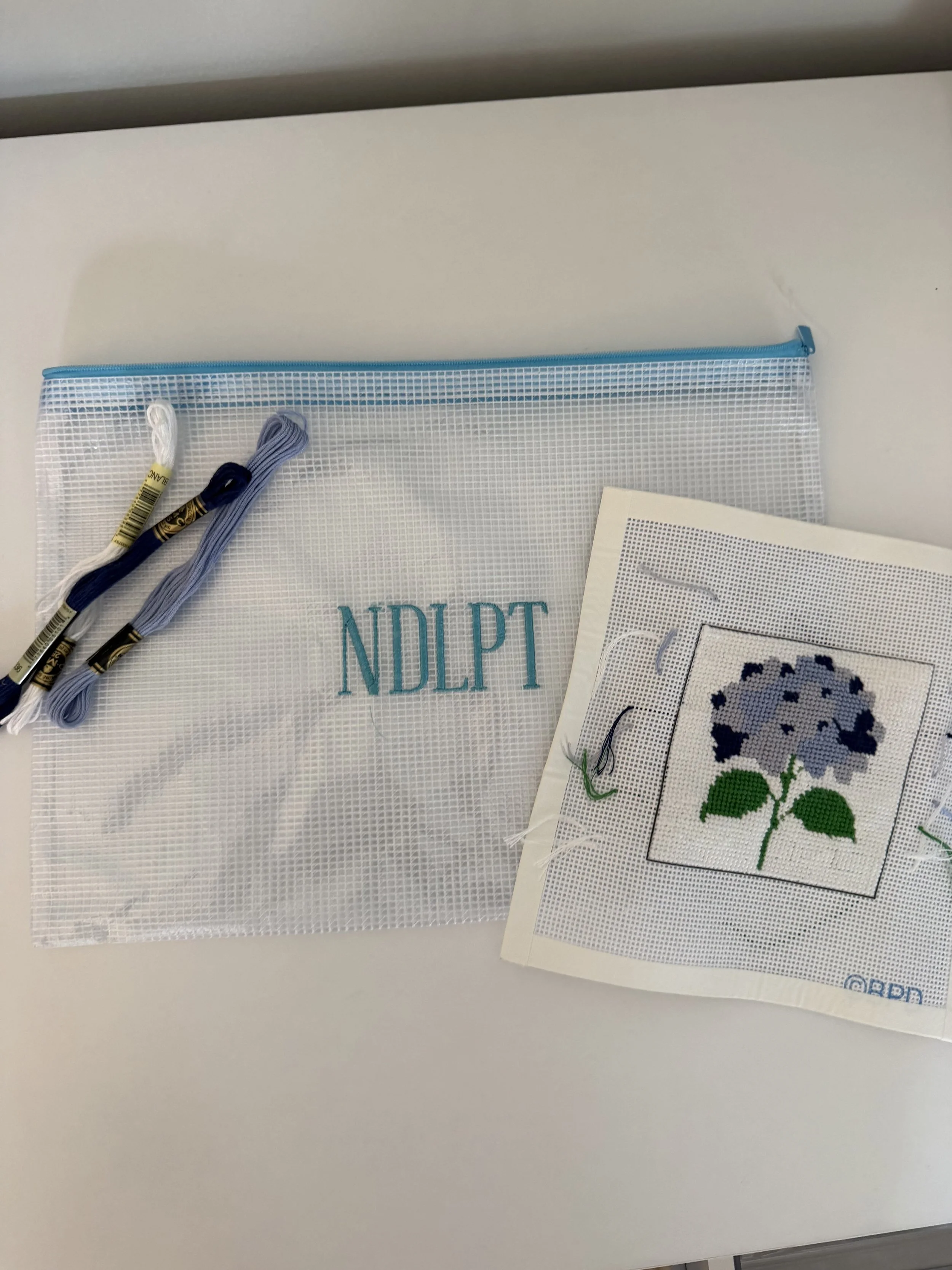 Cross-stitch pattern of a purple flower with green leaves, a plastic zipper bag, embroidery floss in white, blue, and black, and printed cross-stitch pattern sheets.