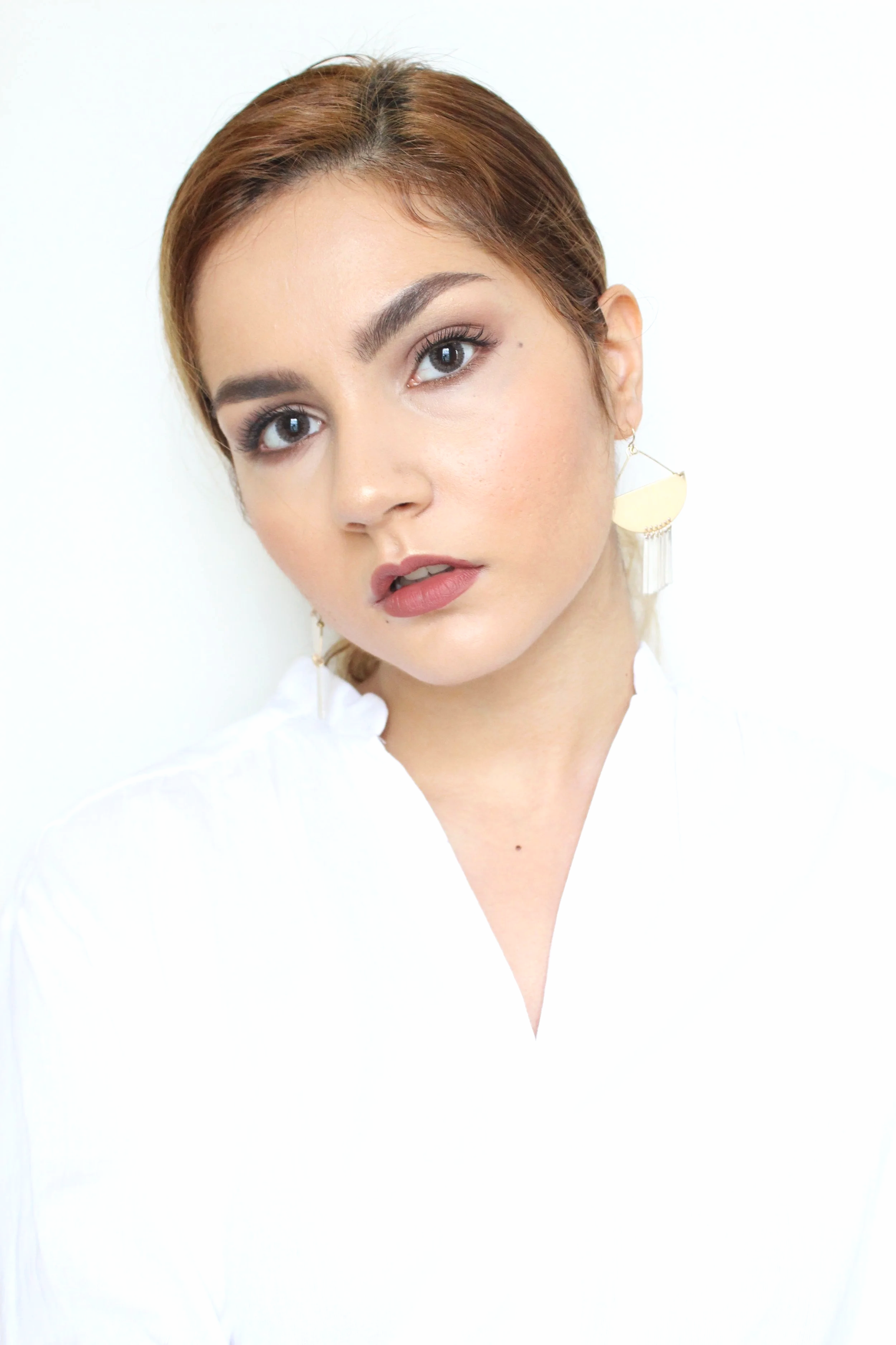 A woman with brown hair styled with a side part, wearing a white top and large cream-colored crescent moon earrings, looking directly at the camera with a neutral expression.