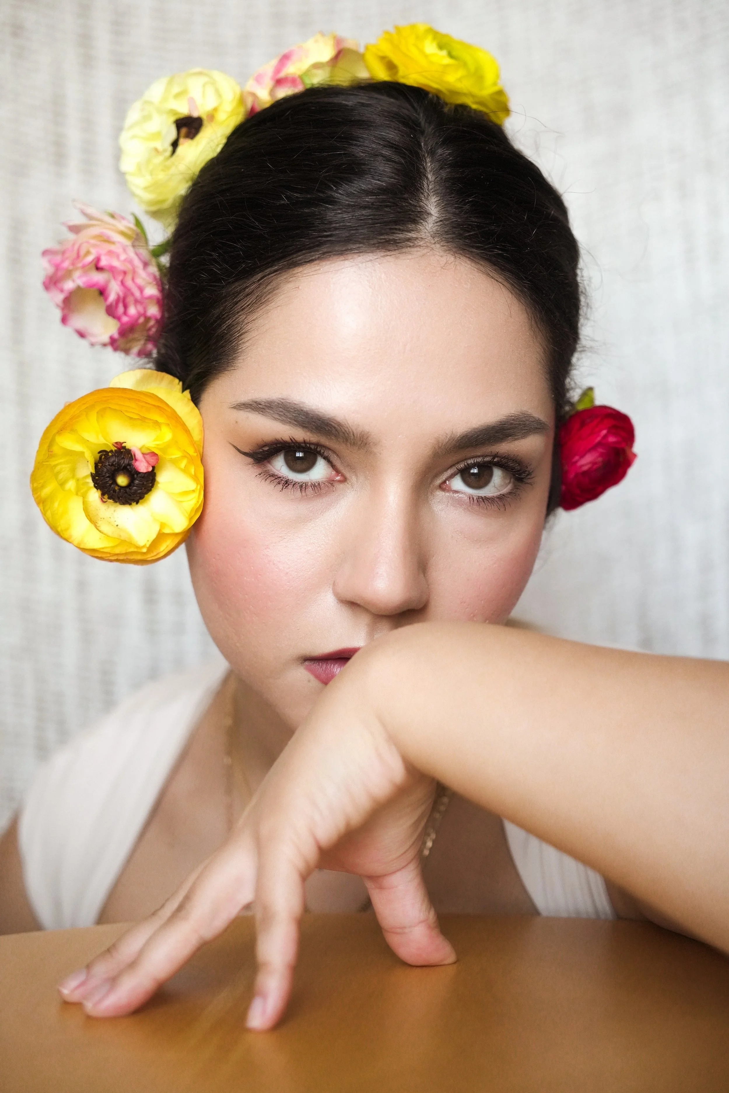 A woman with dark hair and brown eyes, wearing light makeup, rests her chin on her arm and gazes at the camera. She has colorful flowers in her hair, including yellow, pink, and red blooms, and is wearing a white top. The background is a light-colore