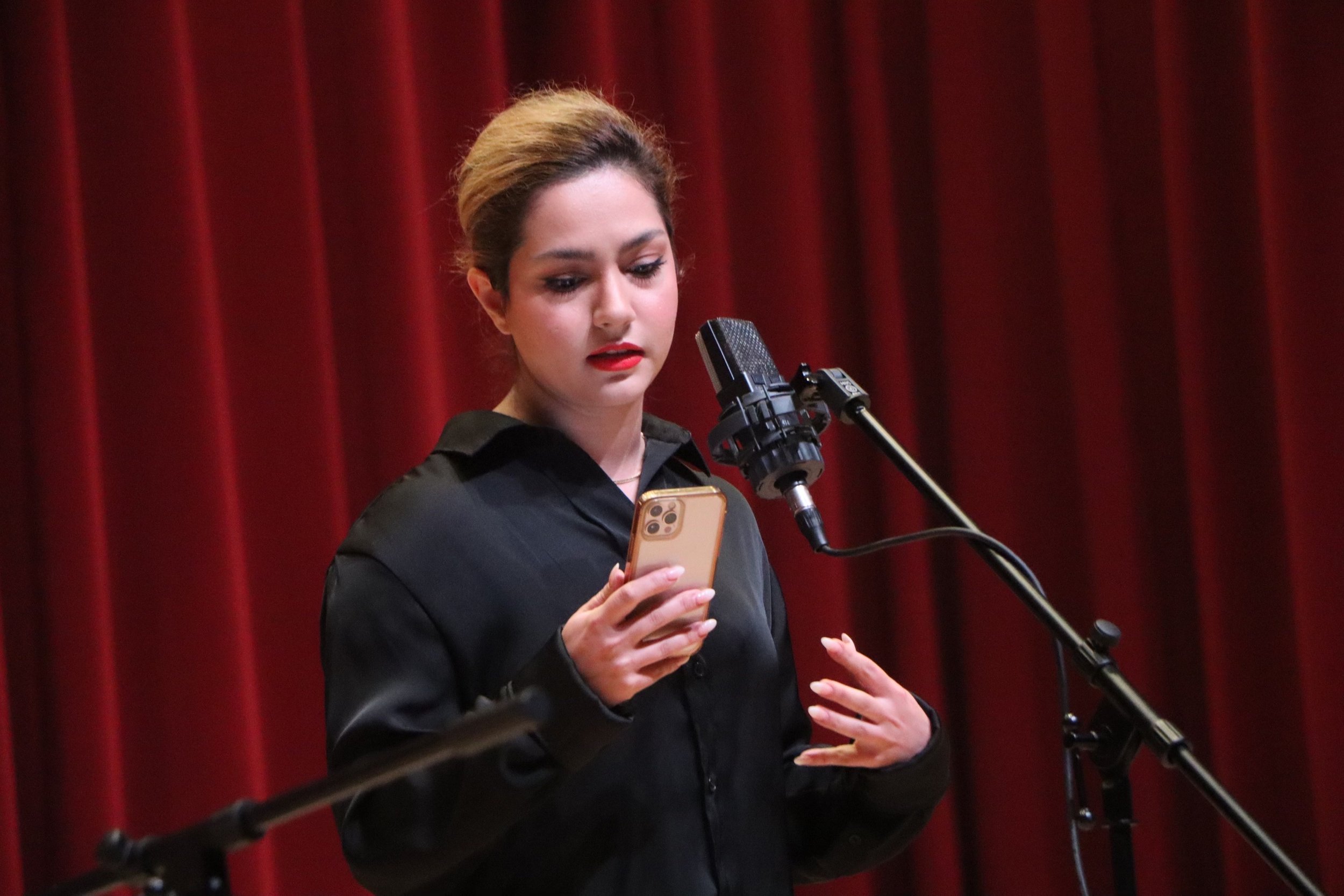 A woman with light brown hair in a bun, wearing a black jacket, speaking or singing into a microphone, holding a smartphone, with a red curtain background.