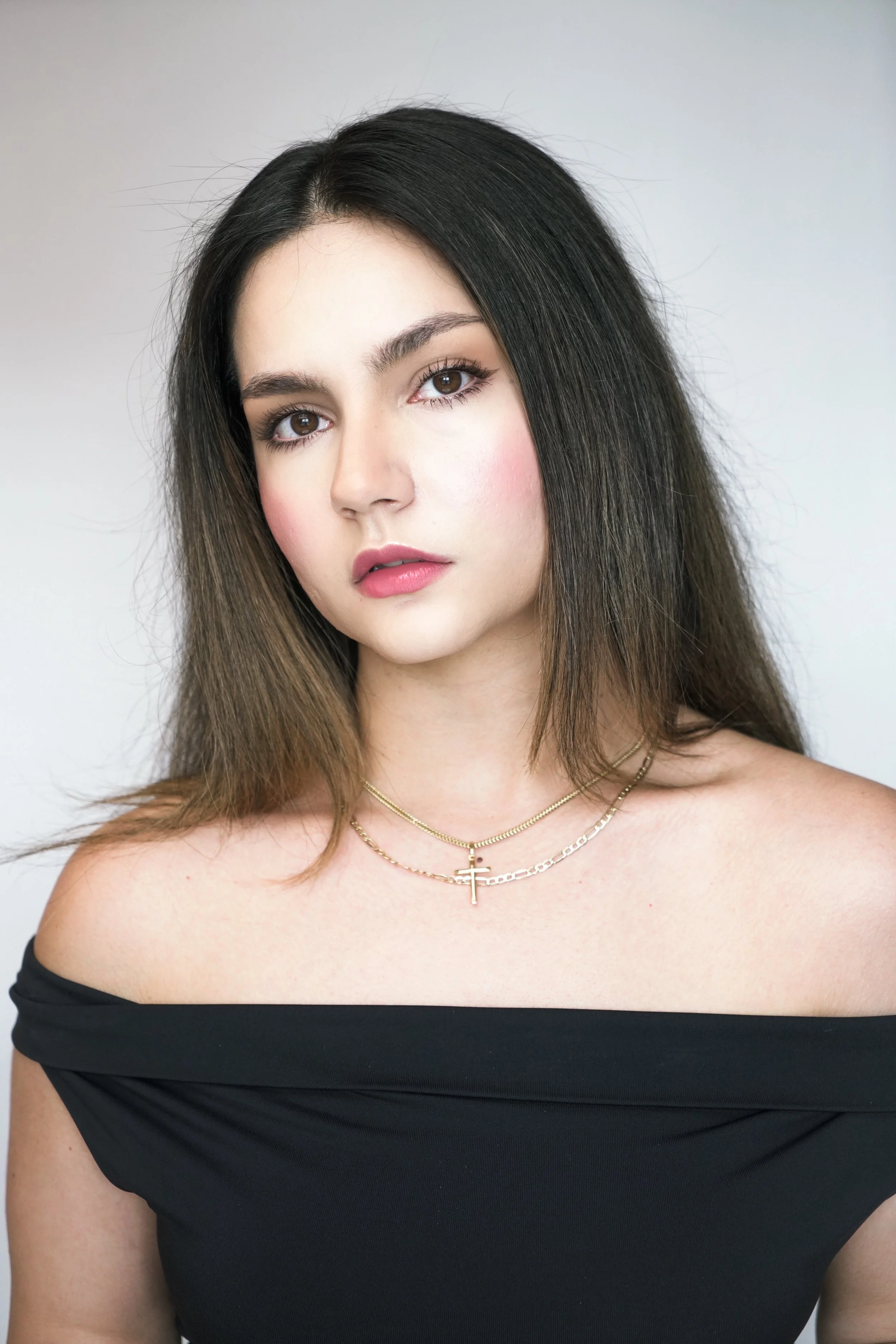Close-up of a young woman with long dark and brown hair, wearing an off-the-shoulder black top and layered gold necklaces, looking directly at the camera against a plain background.