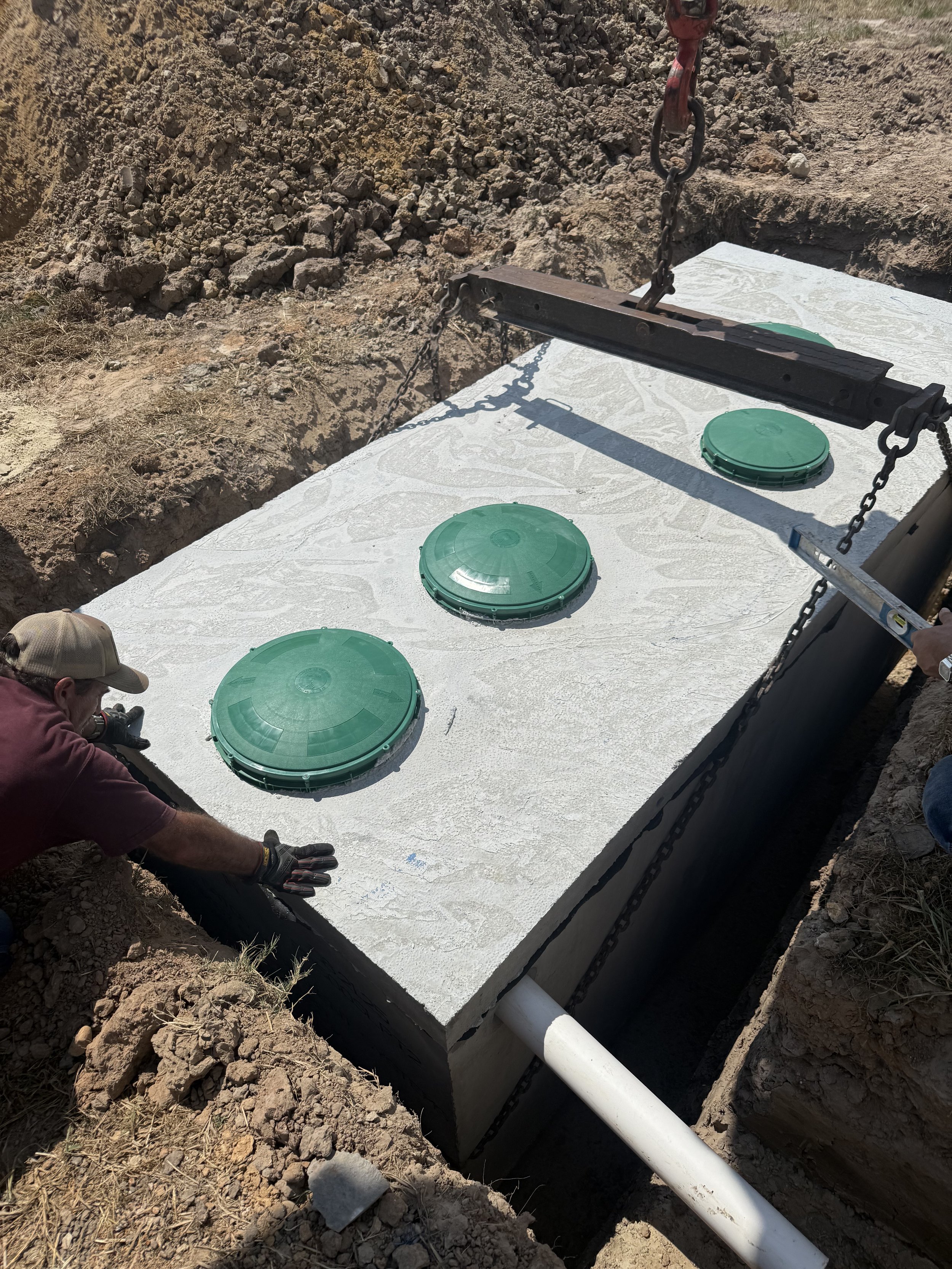 Workers installing a concrete septic tank with four green access covers and a white PVC pipe at a construction site.